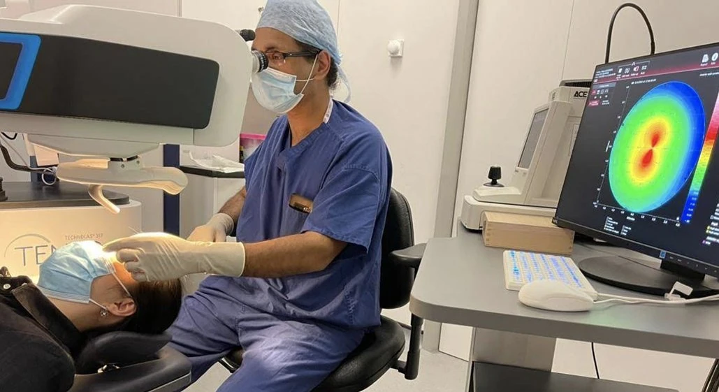 A surgeon in scrubs reviewing a colour corneal topography map on screen while a patient is assessed.