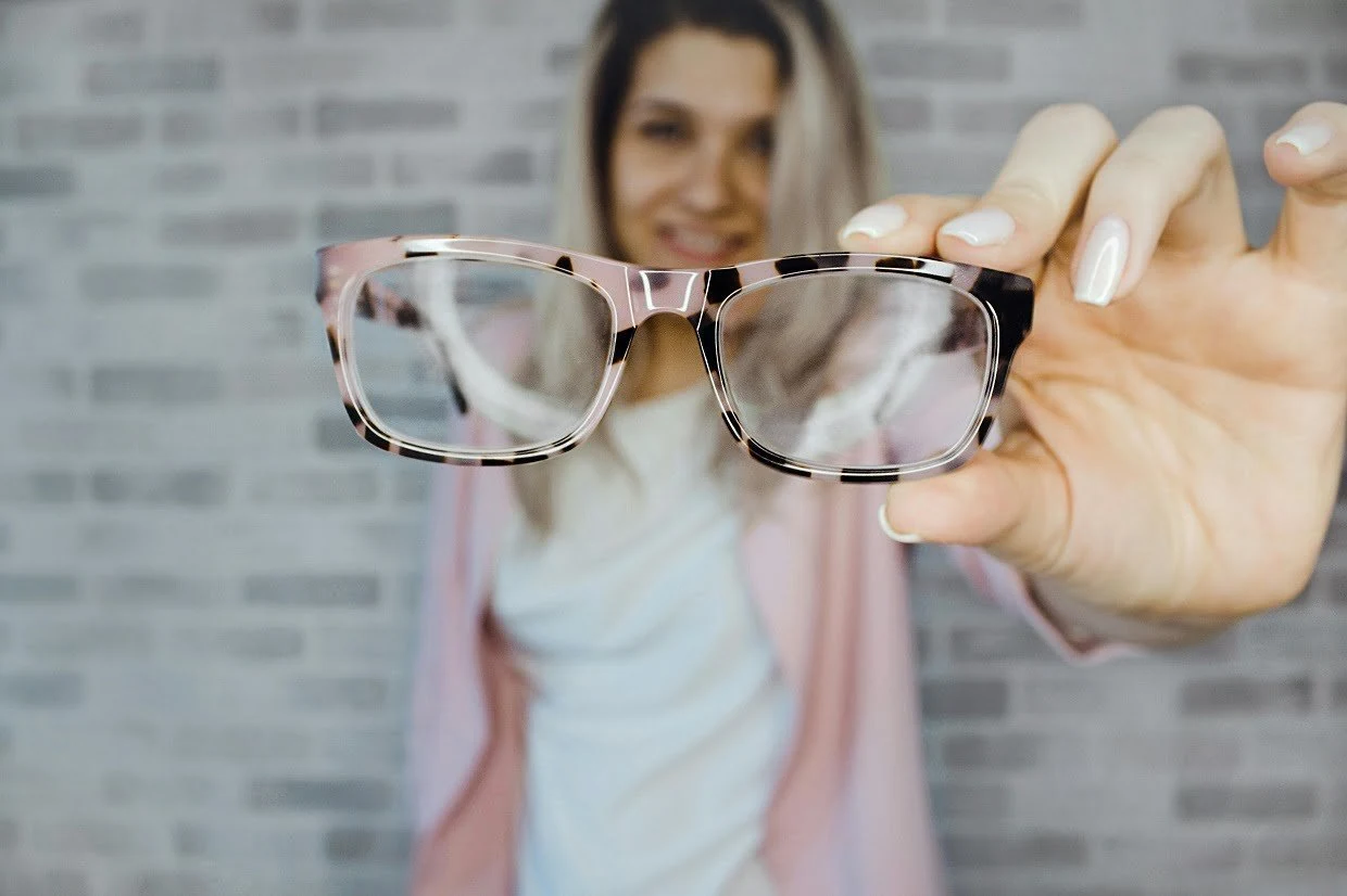 A woman holding her glasses out at arm's length, the lenses sharp and her face blurred behind them.